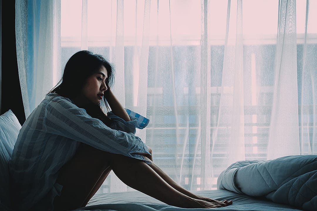 A young adult sitting on a bed and looking pensive near a window in her room.